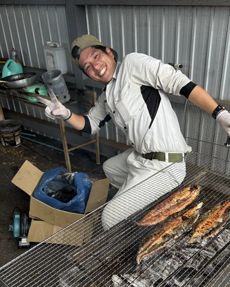 仲村さん。作業小屋で、炭火で川魚を焼きながらピース